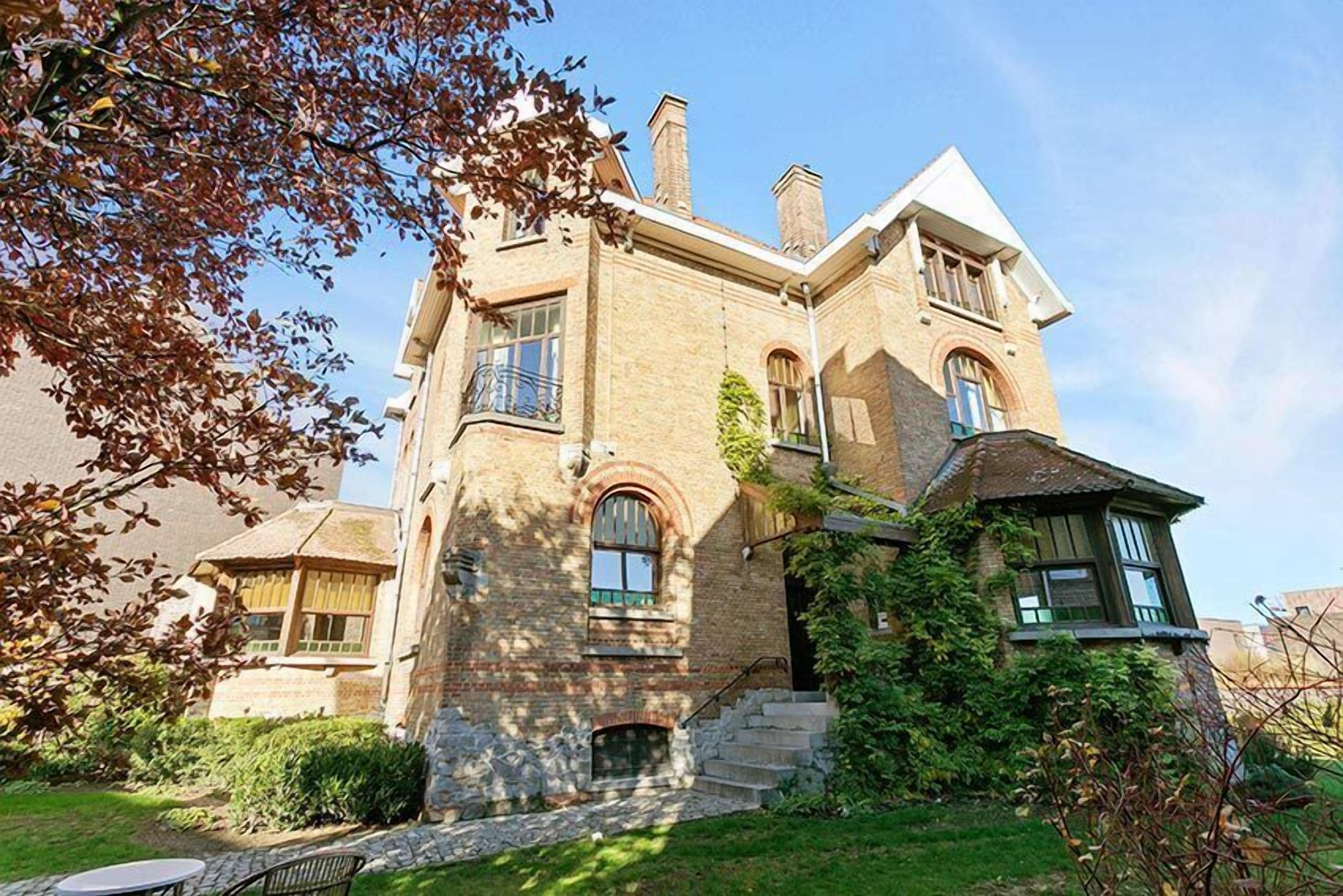 Une maison à deux étages avec une façade en pierre, un porche à l’avant et un petit jardin devant.