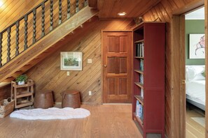 A wooden staircase leading to a loft bedroom with a sloped ceiling, a wooden door, a bookshelf, and a framed picture on the wall.