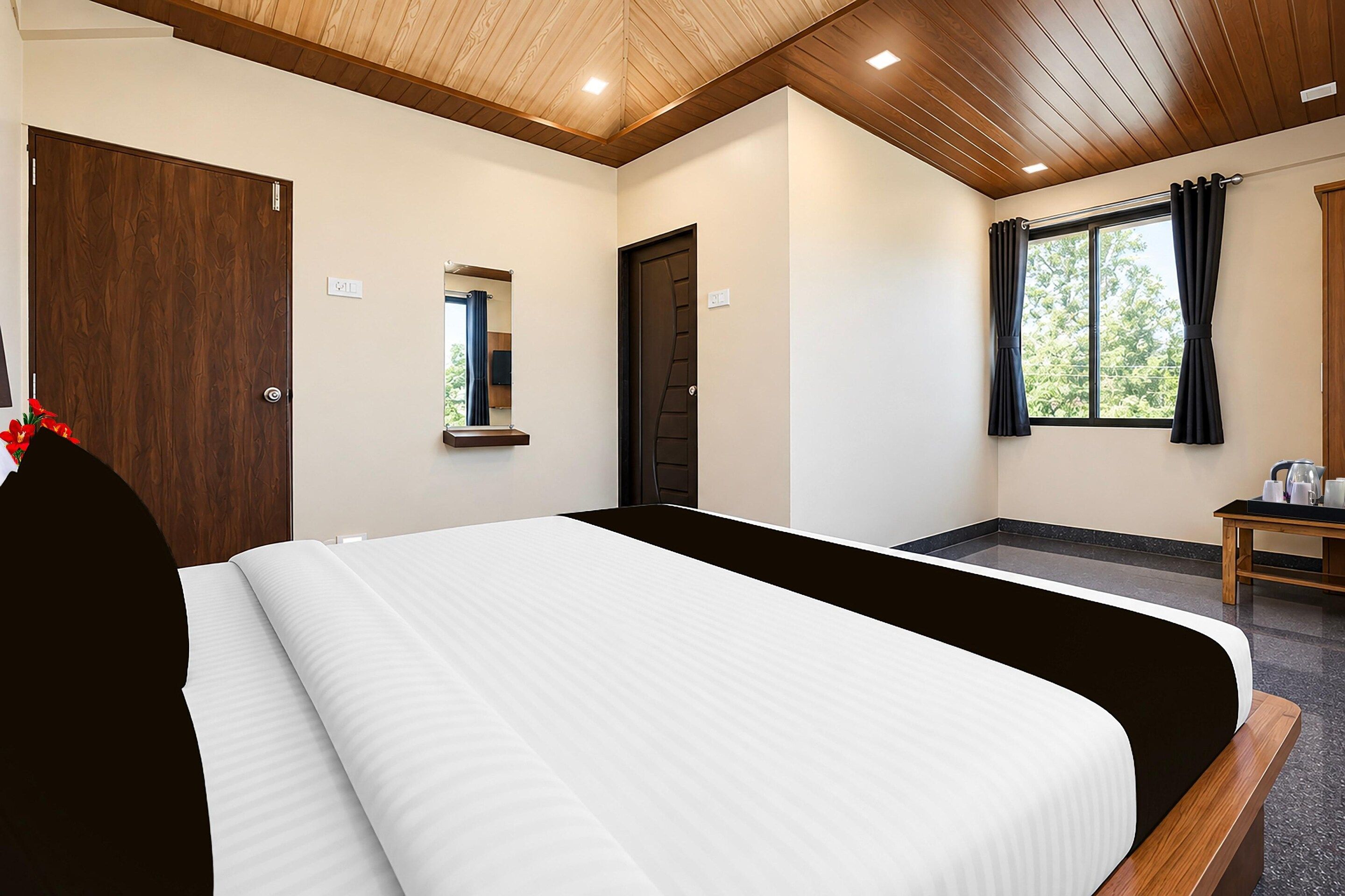 A hotel room with a large bed, a wooden ceiling, a window with curtains, and a small table with a kettle.