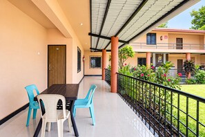 A covered outdoor seating area with a table and chairs, a door, and a view of a building with a balcony.