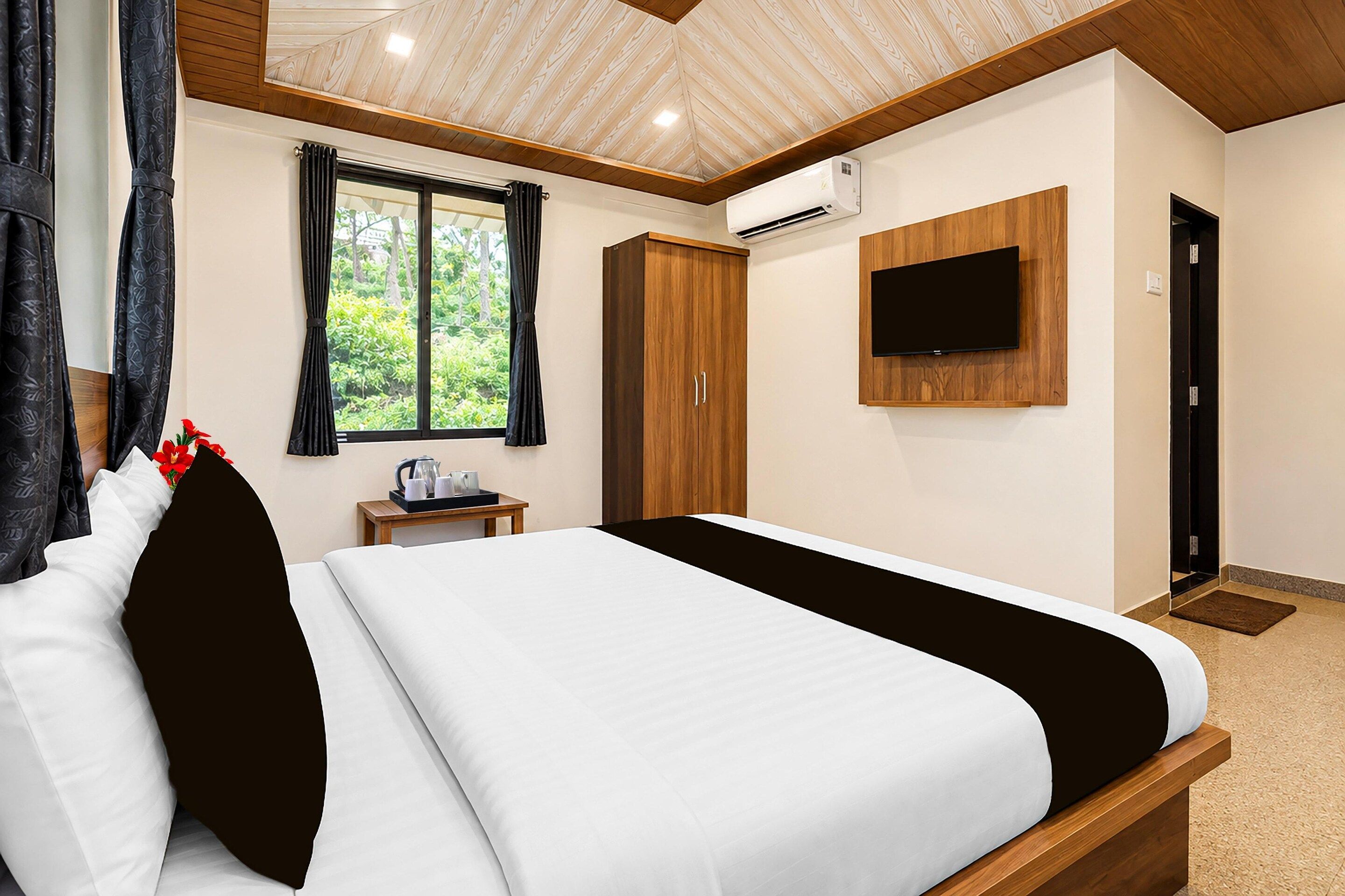 A hotel room with a bed, a TV mounted on the wall, a window with curtains, and an air conditioning unit.