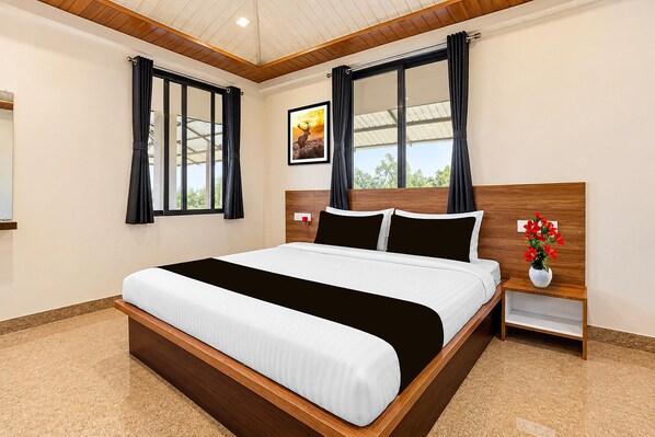 A hotel room with a large bed, wooden headboard, two windows with curtains, a nightstand with a vase of flowers, and a framed picture on the wall. - Classic Jungle Eye (Mahabaleshwar)