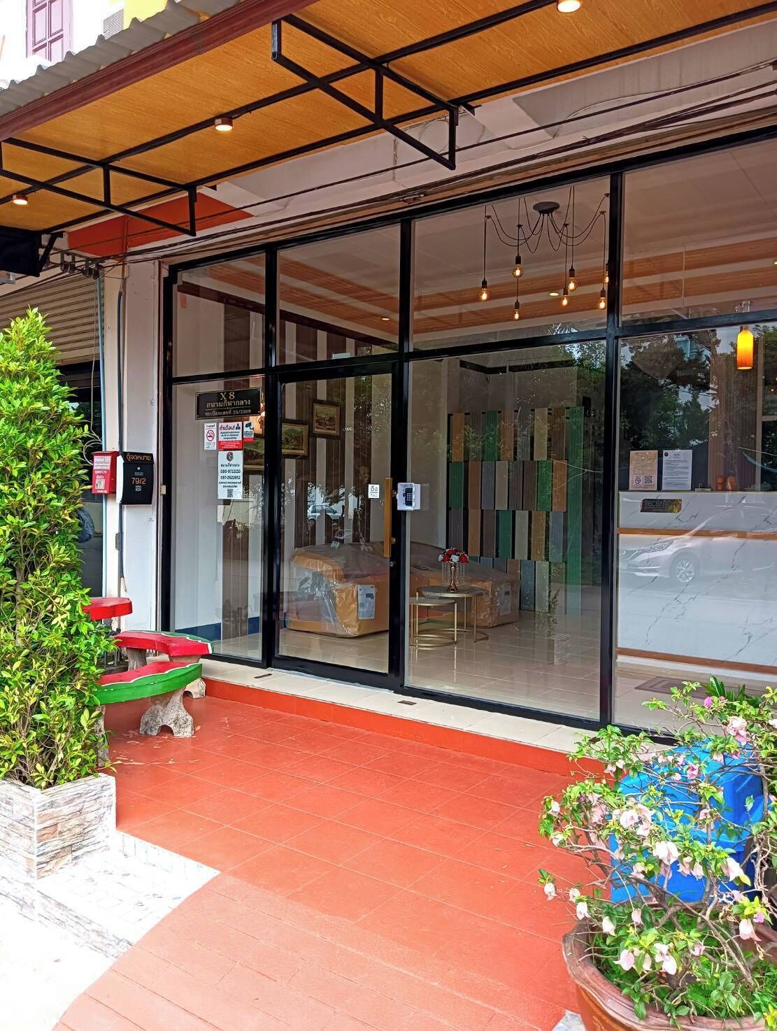 A hotel entrance with a glass facade, a red tiled ground, and a small outdoor seating area with a bench and potted plants.