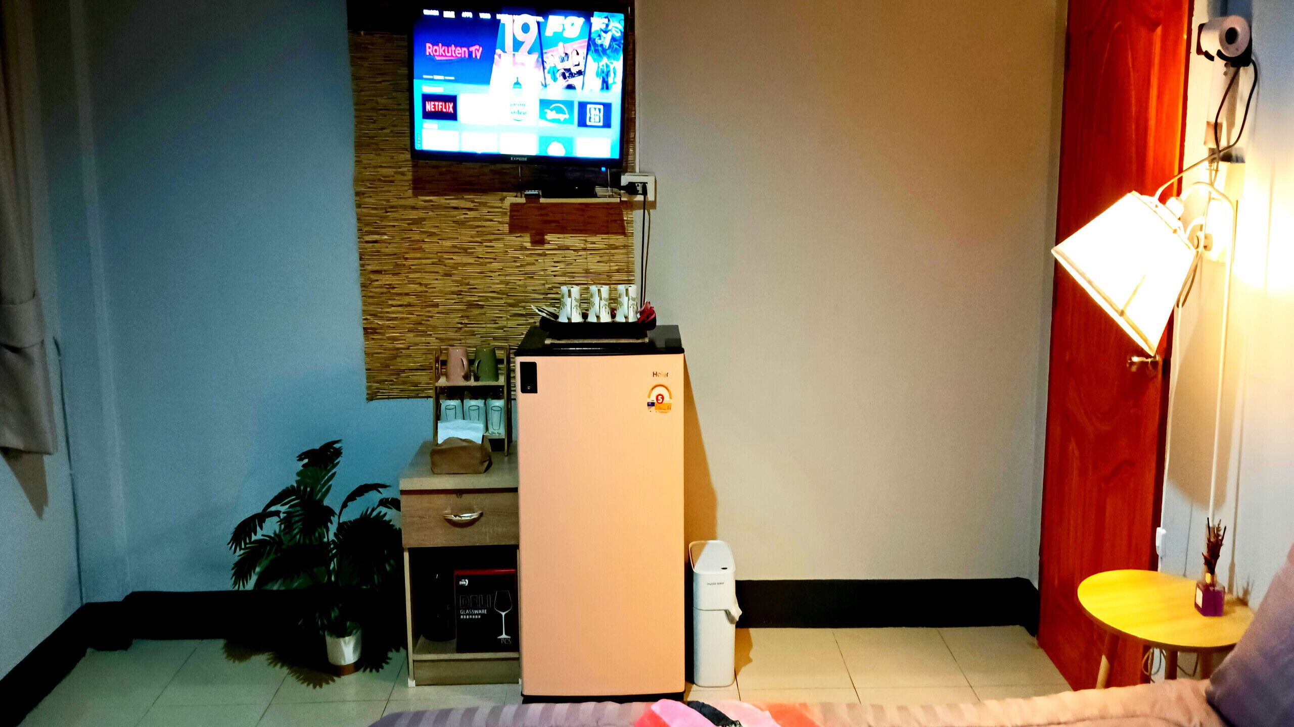 Ein kompakter Wohnbereich mit Wandfernseher, kleinem Kühlschrank, Kaffeemaschine und einem Tisch mit Lampe.