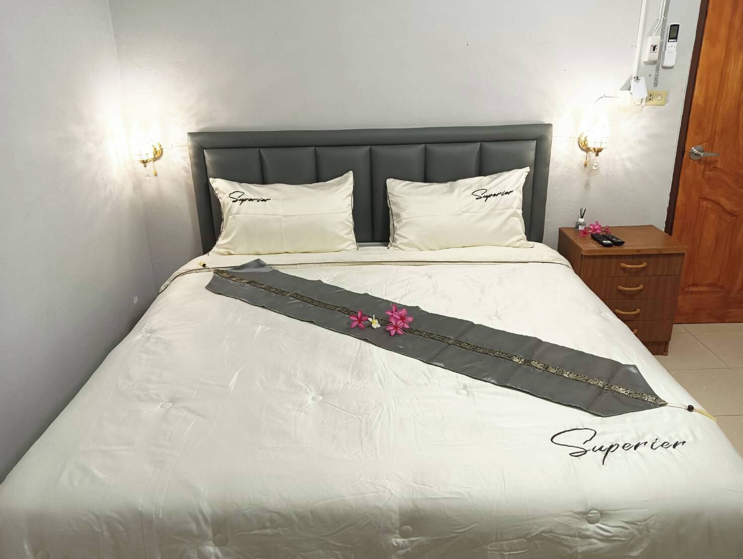 Un lit avec une tête de lit grise, deux oreillers et un couvre-lit blanc orné d’un ruban décoratif.