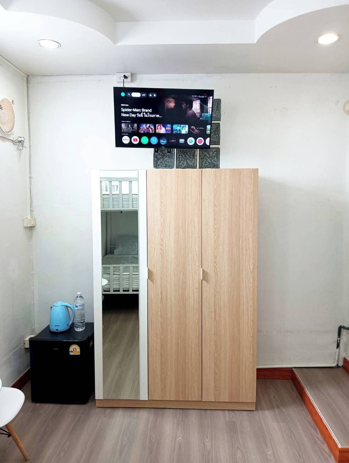 Ein Zimmer mit an der Wand montiertem Fernseher, der einen Film zeigt, einem Kleiderschrank, einem kleinen Tisch mit Wasserkocher und Wasserflasche sowie einem Bett im Hintergrund.