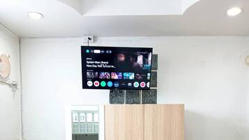 Ein Zimmer mit an der Wand montiertem Fernseher, der einen Film zeigt, einem Kleiderschrank, einem kleinen Tisch mit Wasserkocher und Wasserflasche sowie einem Bett im Hintergrund.