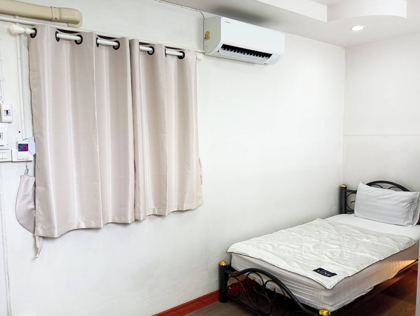 Una cama individual con colchón blanco y estructura metálica negra, pared blanca con equipo de aire acondicionado blanco y cortina color beige.