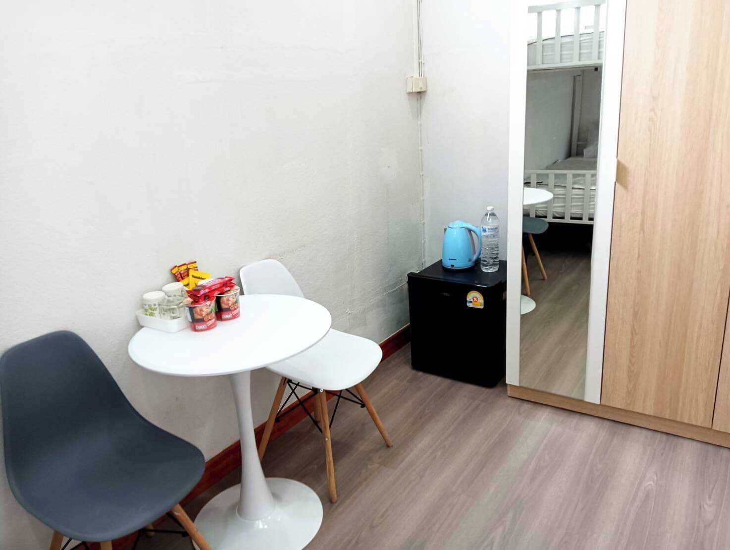 Ein kleines Zimmer mit einem weißen Tisch, zwei Stühlen, einem schwarzen Schrank mit Wasserkocher und Wasserflasche sowie einer Holztür.