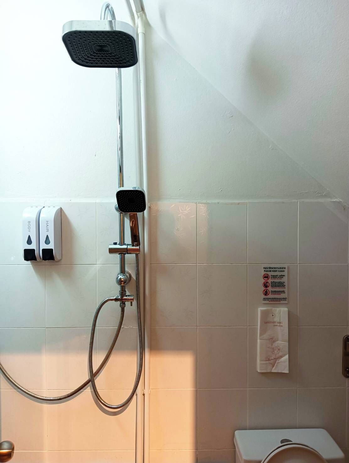 A shower with a handheld showerhead and a handheld scrubber, a soap dispenser, and a sign on the wall.