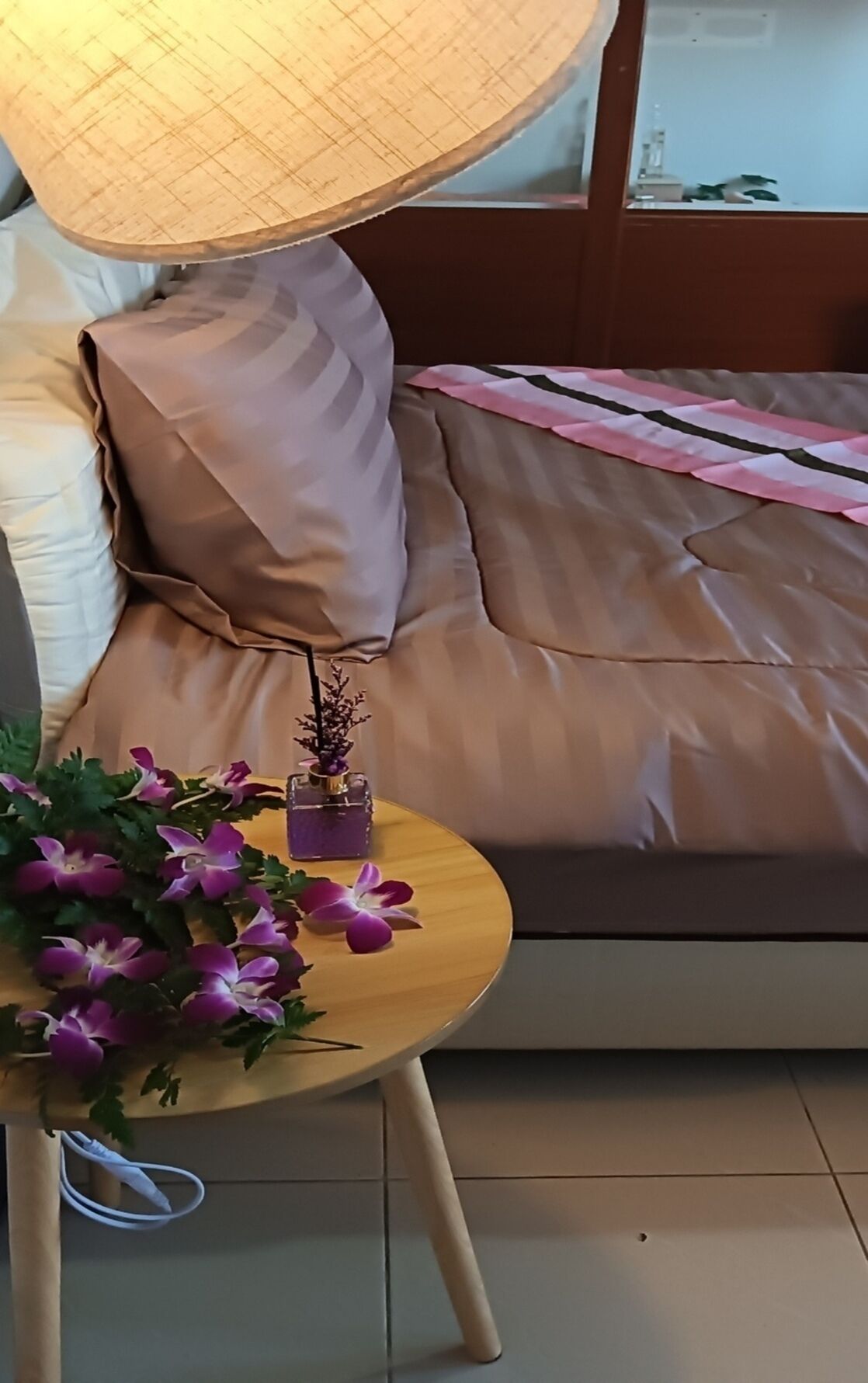 Un lit avec une couverture à rayures, une table de chevet ornée d’un vase de fleurs et une lampe.