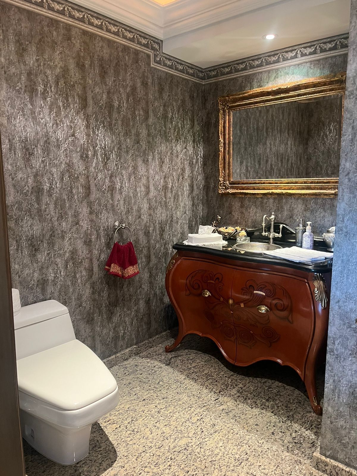A bathroom with a marble floor, a white toilet, a wooden vanity with a gold mirror, and a red towel.