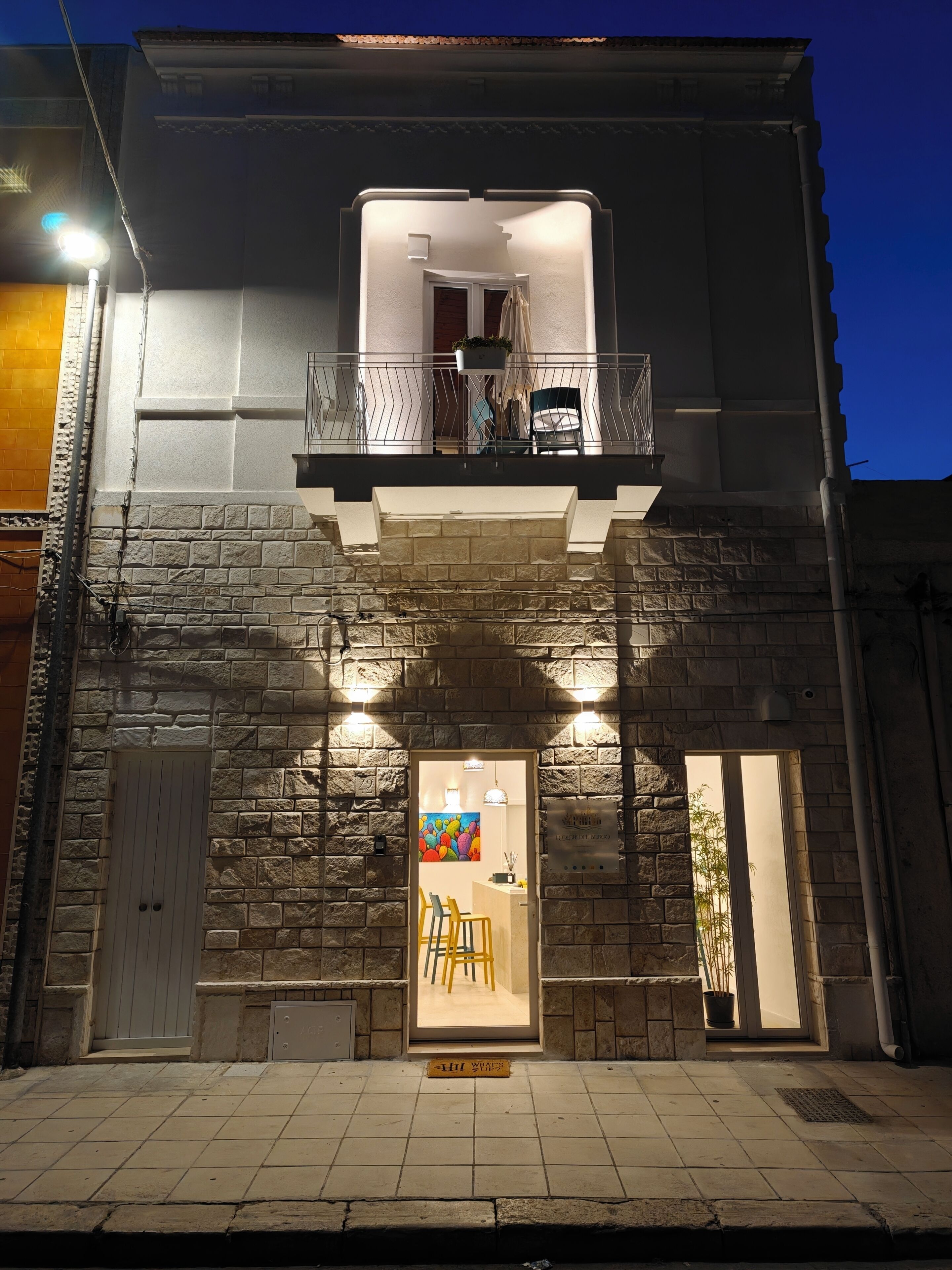 A building with a stone facade, a balcony with a railing, and a door leading to an interior space with a table and chairs.