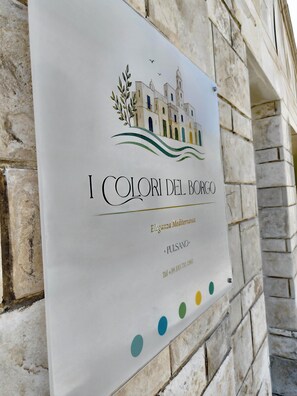 Ein Schild mit einer Illustration eines Gebäudes, dem Namen 'I Colori del Borgo' und dem Standort 'Eleganza Mediterranea - Pulsano'.