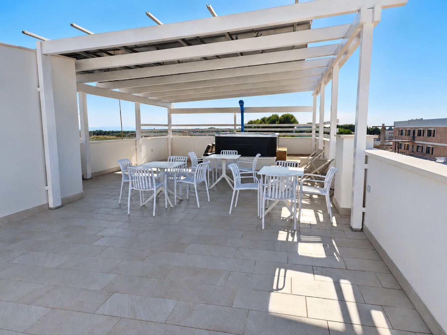 Une terrasse extérieure couverte, avec des chaises et des tables blanches, un barbecue et une vue sur le paysage urbain.