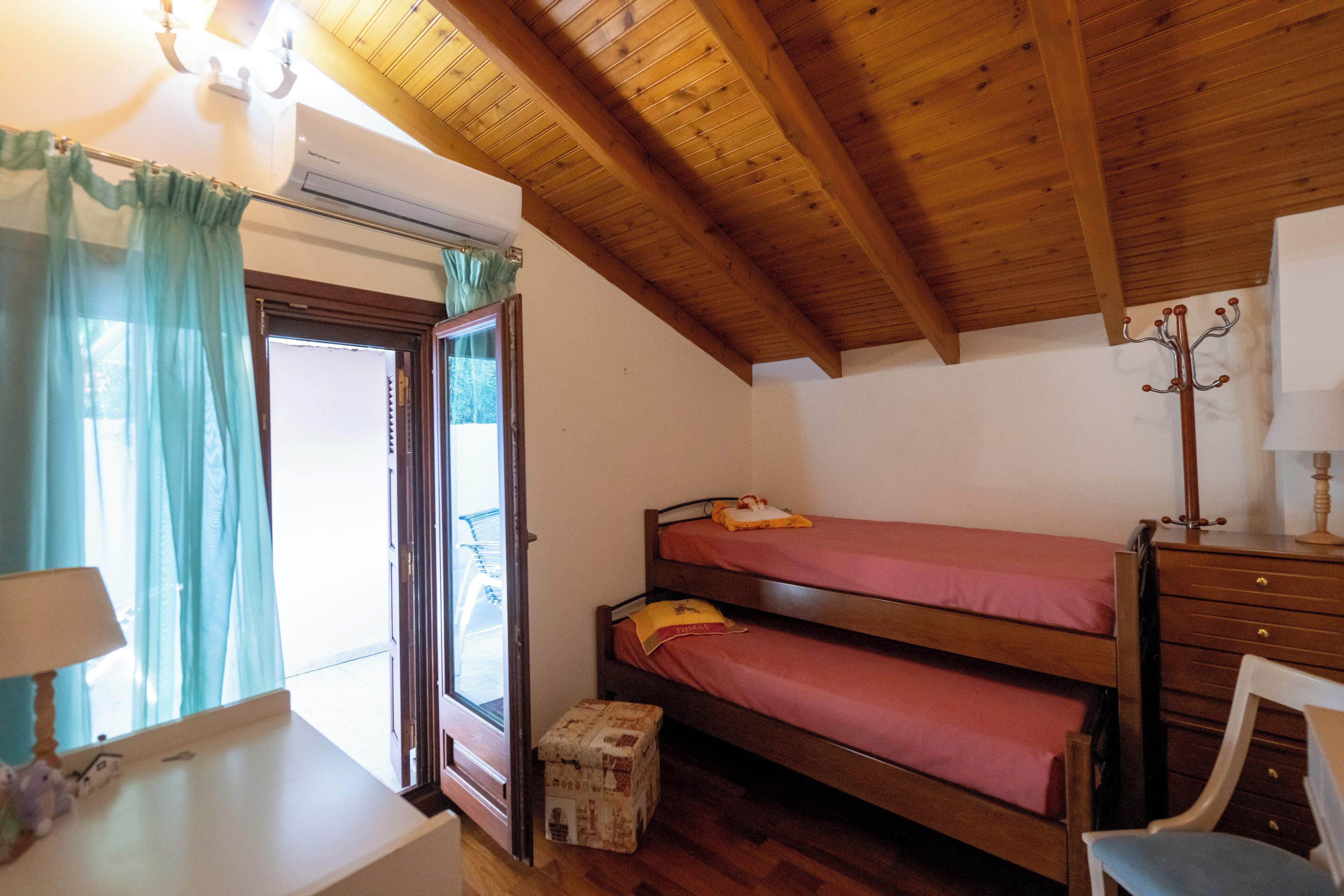A room with a wooden ceiling, a bed with red bedding, a small desk with a lamp, and a window with curtains.