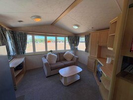 Interior de una caravana compacta con sofá, una mesita, kitchenette y ventanas amplias que dan al exterior.