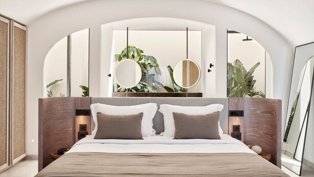A modern bedroom with a large bed, two bedside tables, a mirror, and a view of greenery. - Elyrion Suite, Divine Sea Views, Private Pool, Furnished Terrace, Sanrotini (Oia, Santorini)