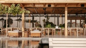 An outdoor seating area with wooden tables and chairs, surrounded by potted plants and a pool. - Elyrion Suite, Divine Sea Views, Private Pool, Furnished Terrace, Sanrotini (Oia, Santorini)