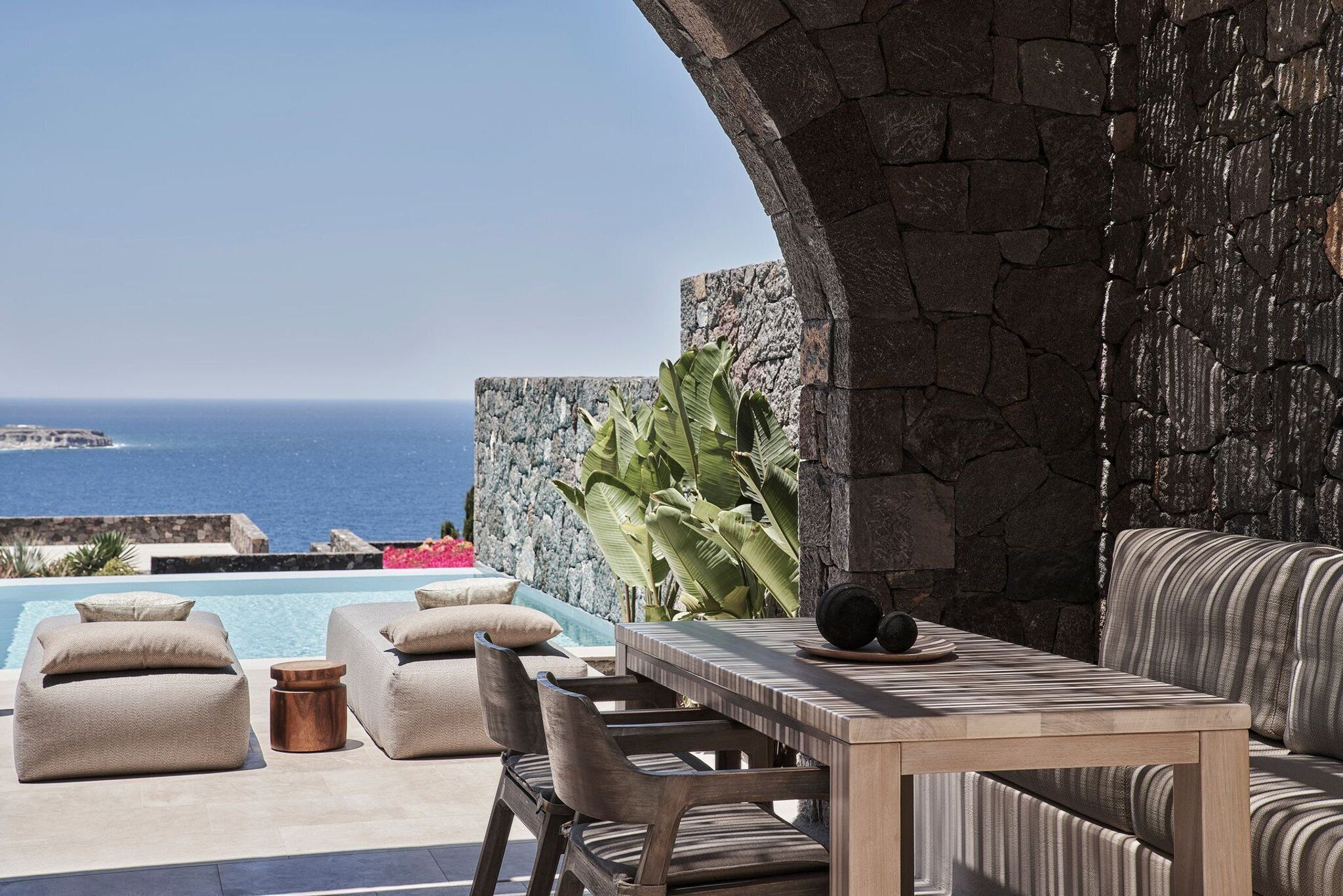 An outdoor seating area with a pool, stone wall, and a view of the sea.