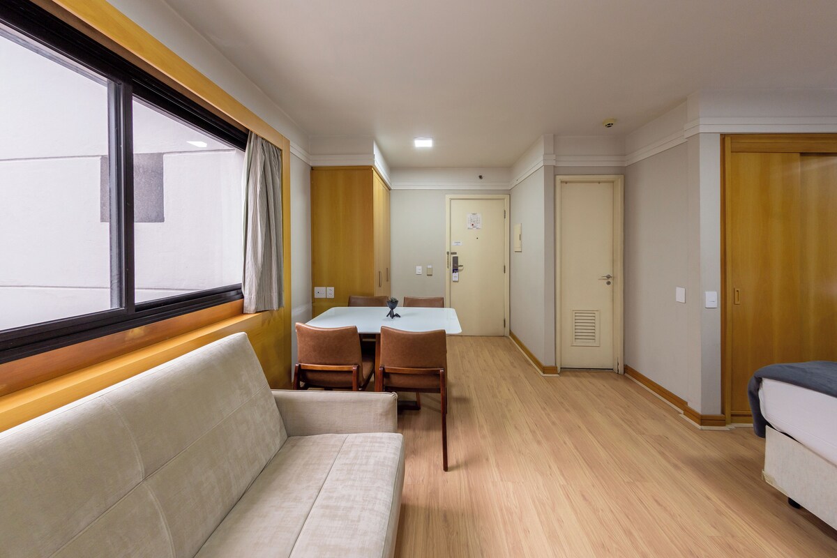 A hotel room with a sofa, a dining table with chairs, a small kitchen area, and a door leading to another room.