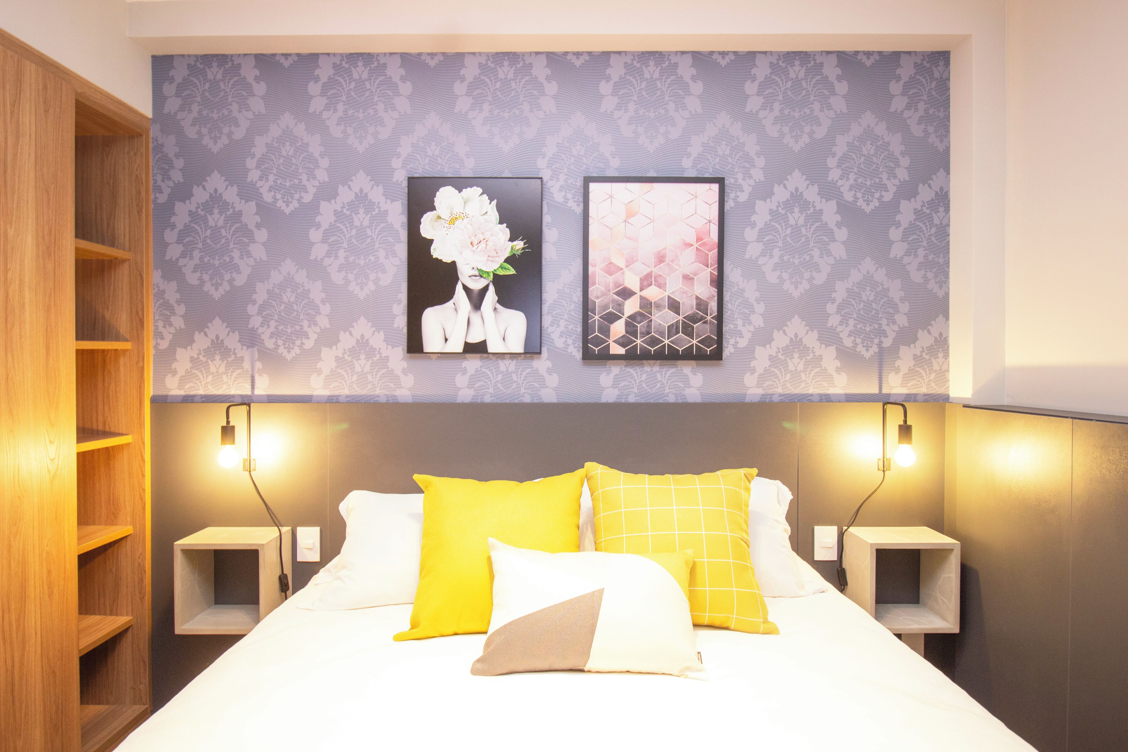 A hotel room with a bed, two bedside tables, a nightstand, a wooden wardrobe, and two wall-mounted artworks.