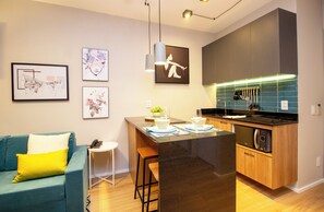Een moderne keuken met een centraal eiland, barkrukken en een eethoek. Er is een kitchenette met een magnetron en een kookplaat. De kamer heeft een blauwe bank met een gele kussens en een klein bijzettafeltje.