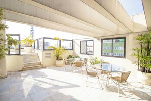 Pátio iluminado pelo sol, com mesa de vidro e cadeiras de vime, rodeado por plantas em vasos e vista para edifícios.
