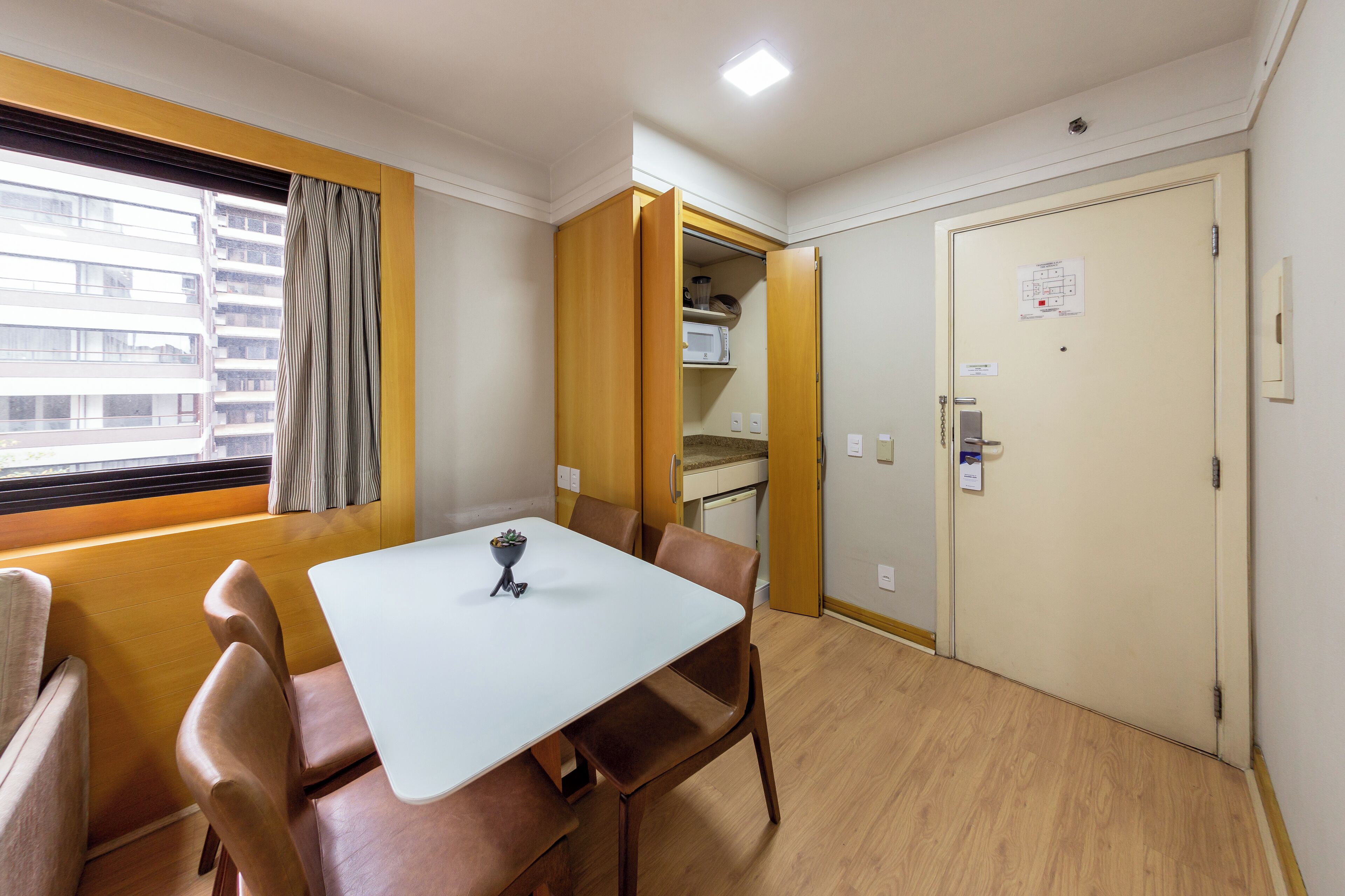 Quarto de hotel com mesa de jantar, cadeiras, janela com cortinas, cozinha compacta e porta com fechadura.