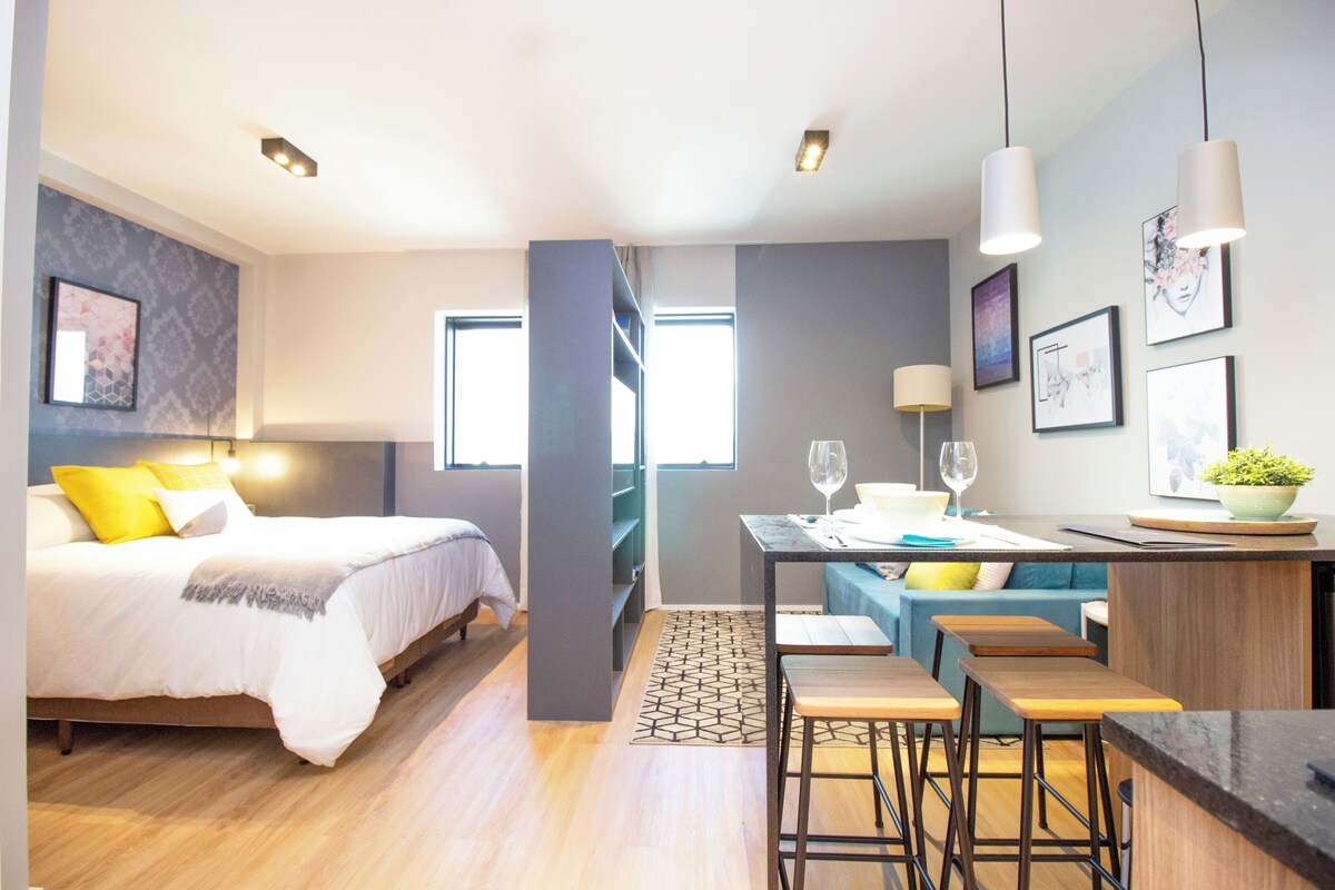 A modern hotel room with a bed, a dining area with a table and chairs, and a kitchen counter.