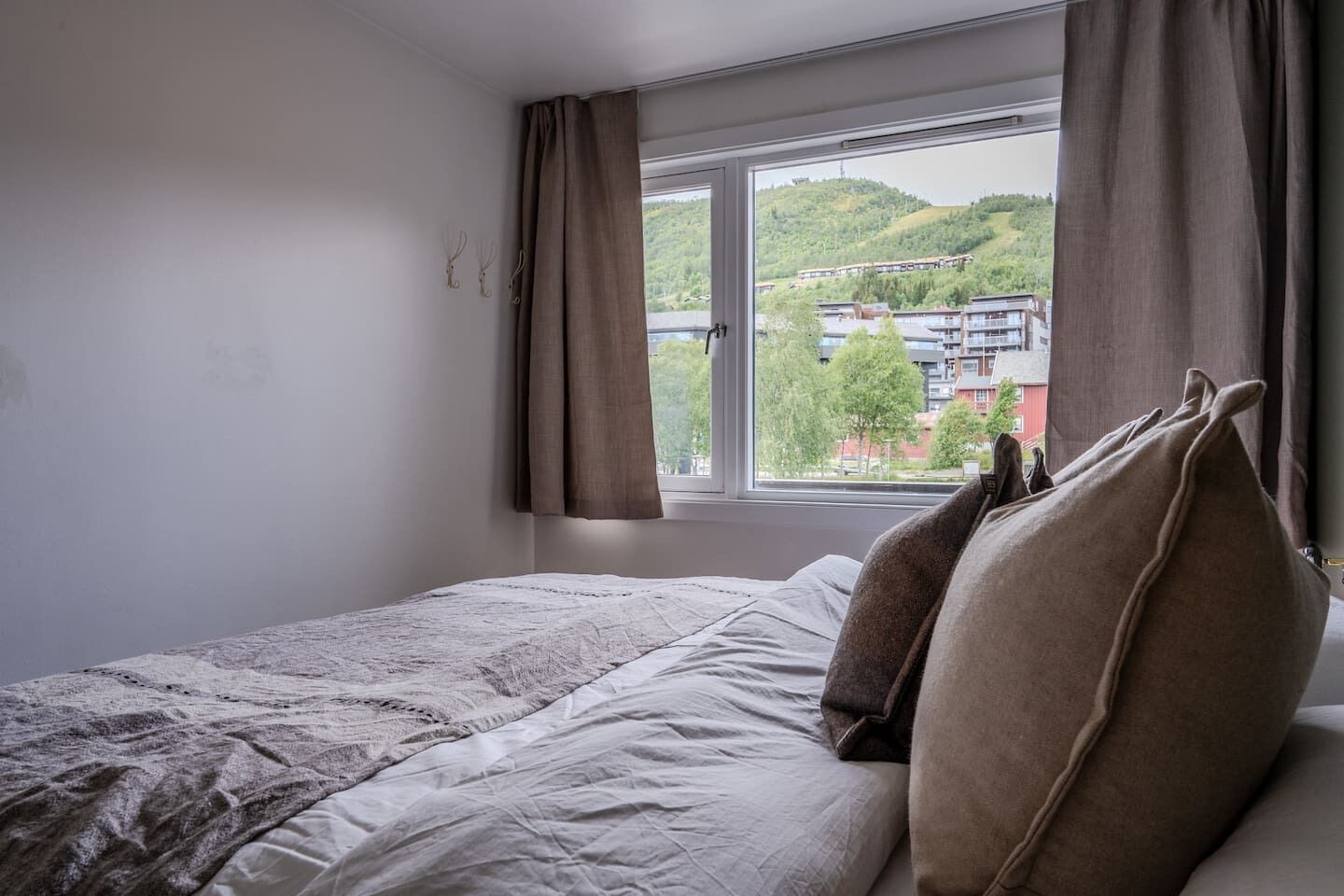 A bedroom with a bed, a window with curtains, and a view of buildings and greenery outside.