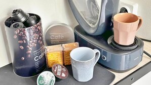 A coffee maker, a mug, a container with coffee beans, a box of biscuits, and a container with a lid.