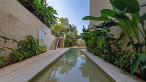 Piscina con borde de azulejos, rodeada de abundante vegetación y una escalera que lleva a un área cubierta.
