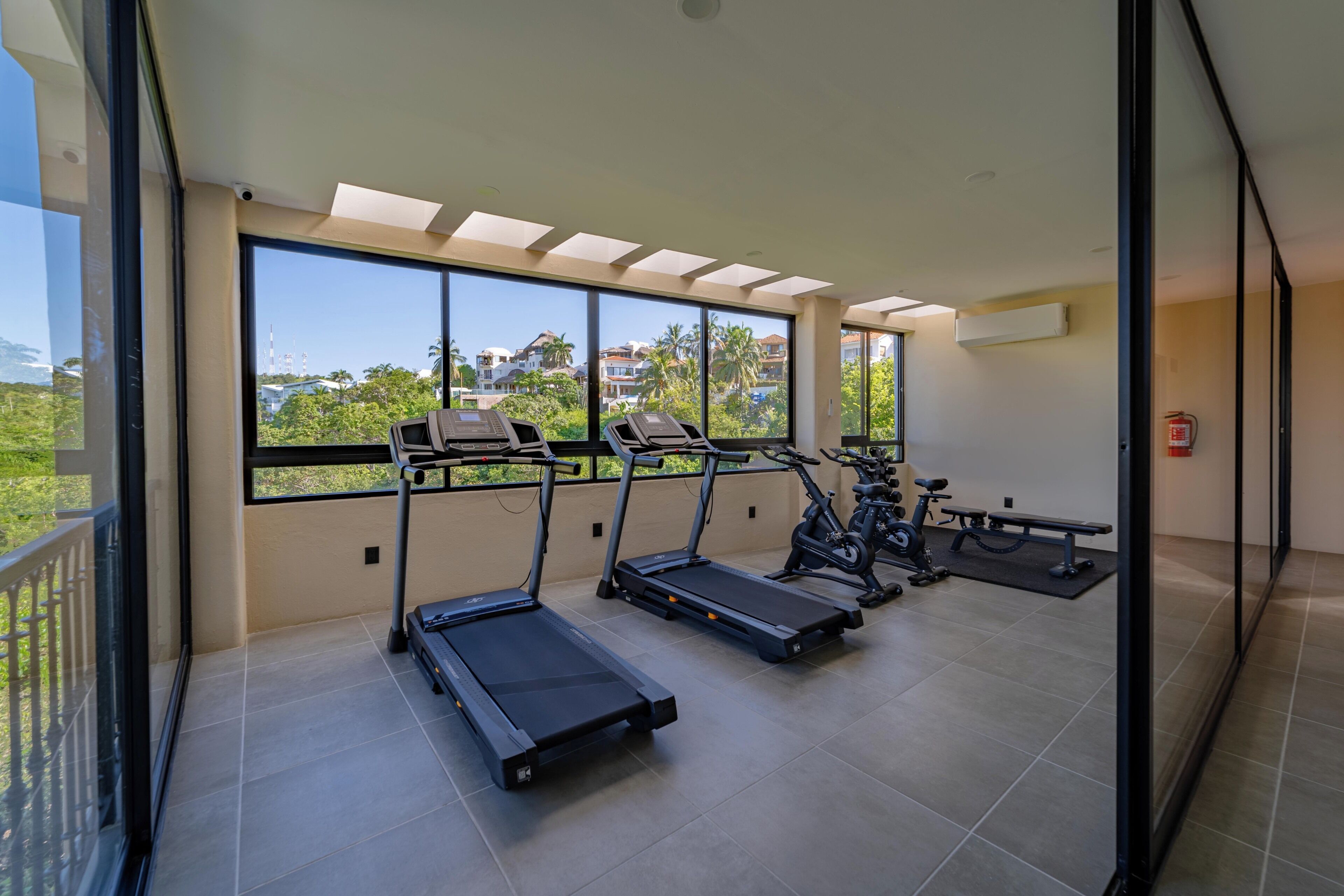 Une salle de sport équipée de tapis de course, d’un vélo d’exercice et d’un banc. De grandes fenêtres offrent une vue sur les bâtiments et les arbres.