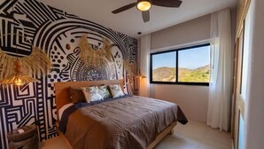 Un dormitorio con una cama grande, un ventilador de techo, una ventana con vistas al paisaje y una pared con un diseño geométrico.