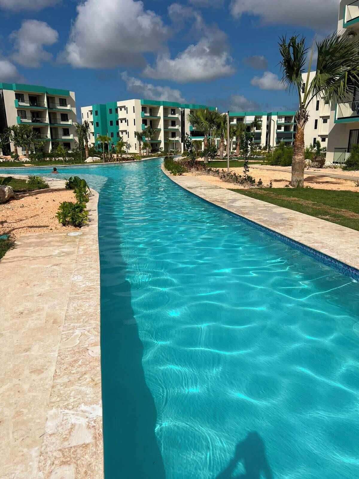 A swimming pool with clear blue water, surrounded by a landscaped area with palm trees and a building complex in the background.