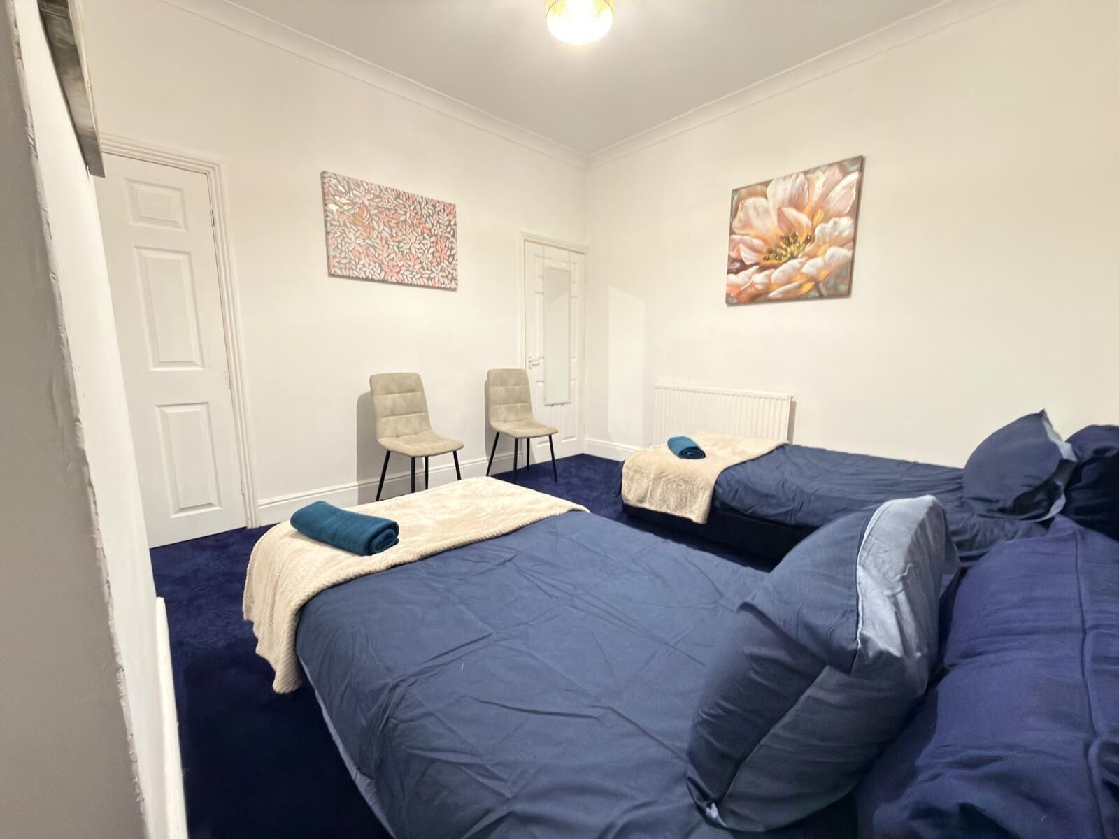 Hotellihuone, jossa on kaksi sänkyä, valkoinen ovi, kaksi tuolia ja kaksi seinätaideteosta.