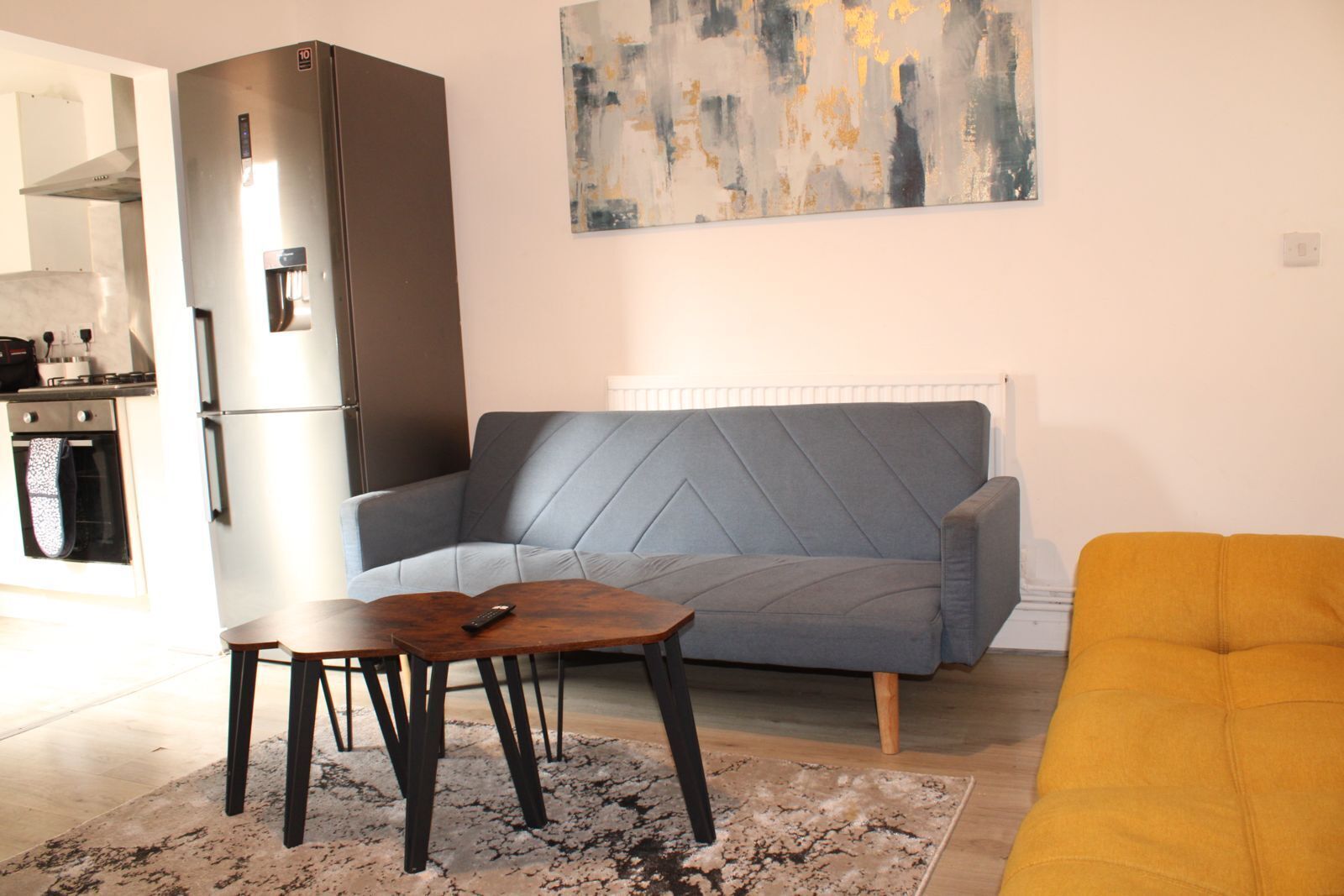 A living room with a grey sofa, a wooden coffee table, a yellow armchair, a kitchen area with a refrigerator, and a painting on the wall.