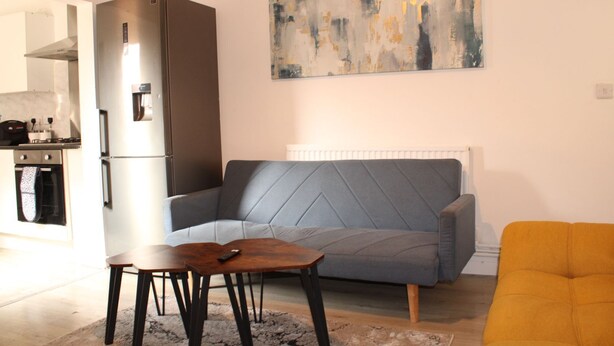 A living room with a grey sofa, a wooden coffee table, and a yellow armchair. A modern kitchen with a stainless steel refrigerator is visible in the background. - Spacious 3BR Doncaster Home Near Racecourse, Shopping & Attractions (Doncaster)