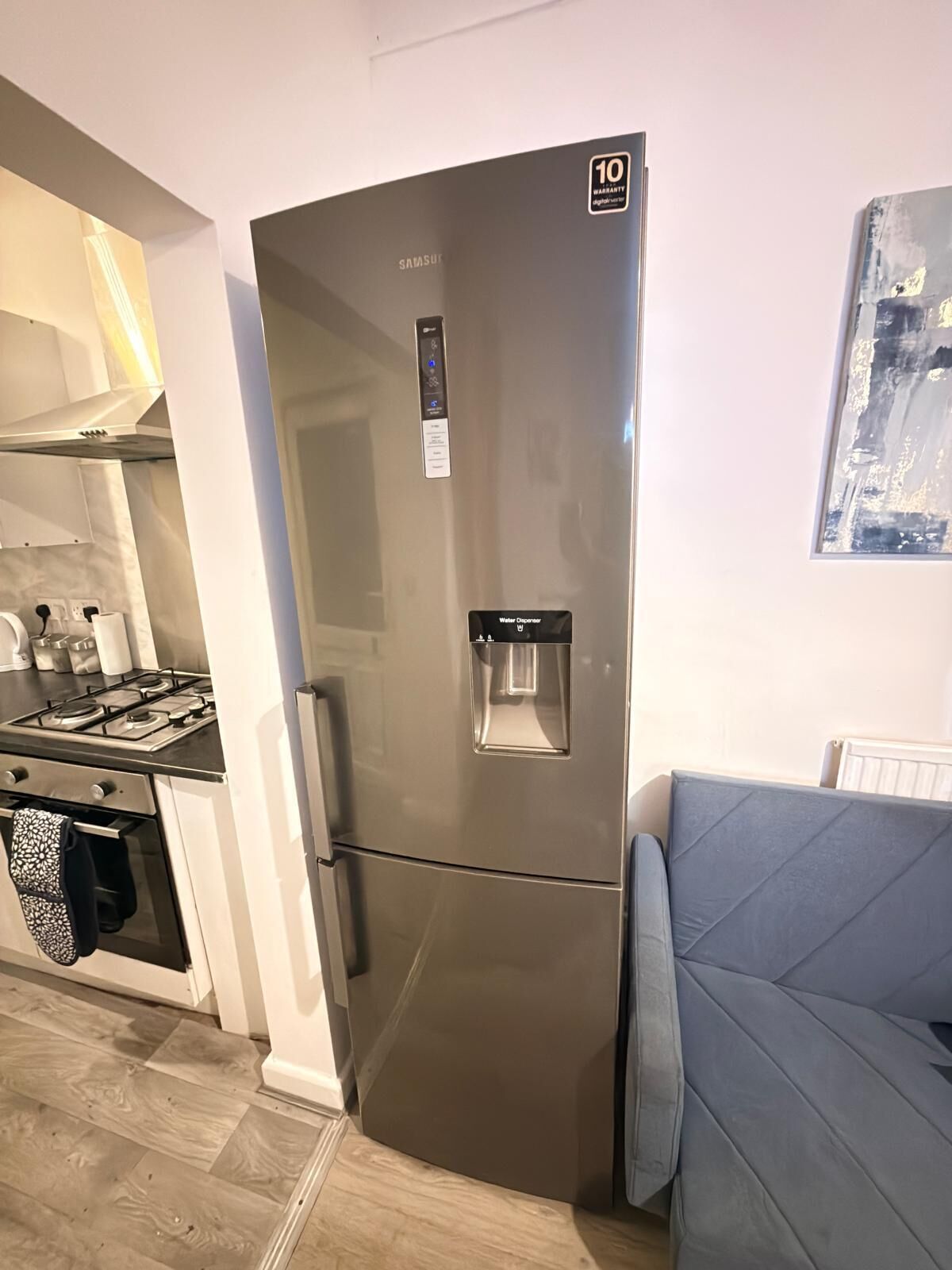 A stainless steel refrigerator with a water dispenser, a stove, and a blue couch in a modern kitchen.
