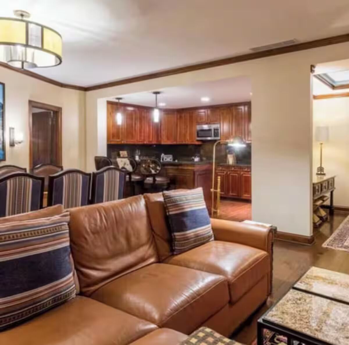A living room with a brown leather sofa, a dining table with chairs, and a kitchen area with wooden cabinets and a microwave.