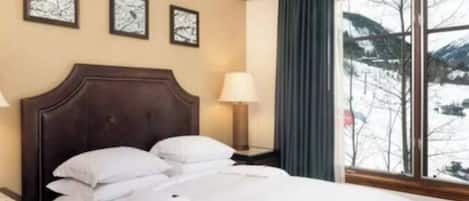 A hotel room with a bed, a nightstand with a lamp, and a window with a view of snow-covered mountains.
