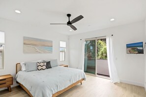 A bedroom with a bed, a nightstand, a ceiling fan, a large window with curtains, and a painting on the wall. - Beacons Beach Retreat | Steps from the Sand (Encinitas)