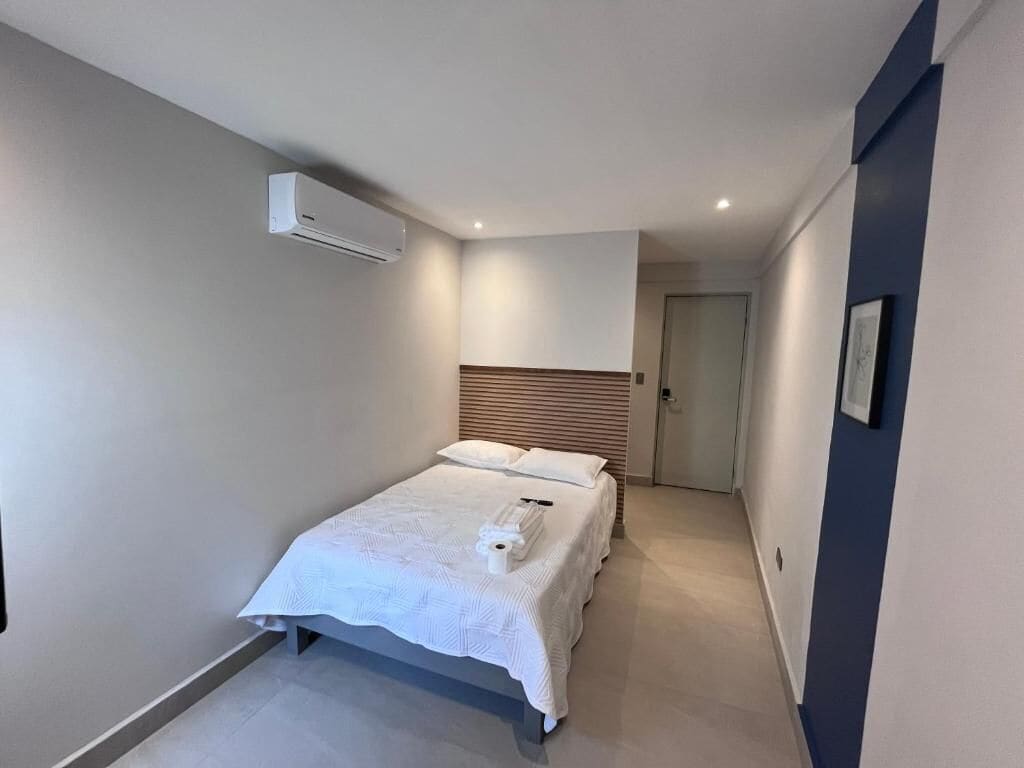 Una habitación de hotel moderna con una cama, una mesita de noche, aire acondicionado en la pared y una puerta que da a otra habitación.