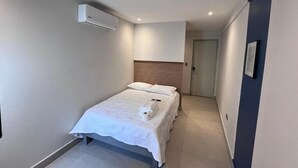 Una habitación de hotel moderna con una cama, una mesita de noche, aire acondicionado en la pared y una puerta que da a otra habitación.
