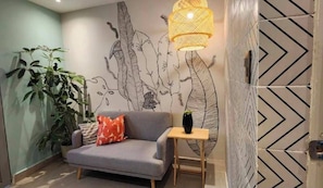 Een moderne woonkamer met een grijze bank, een klein houten bijzettafeltje, een wandlamp en een grote kamerplant.