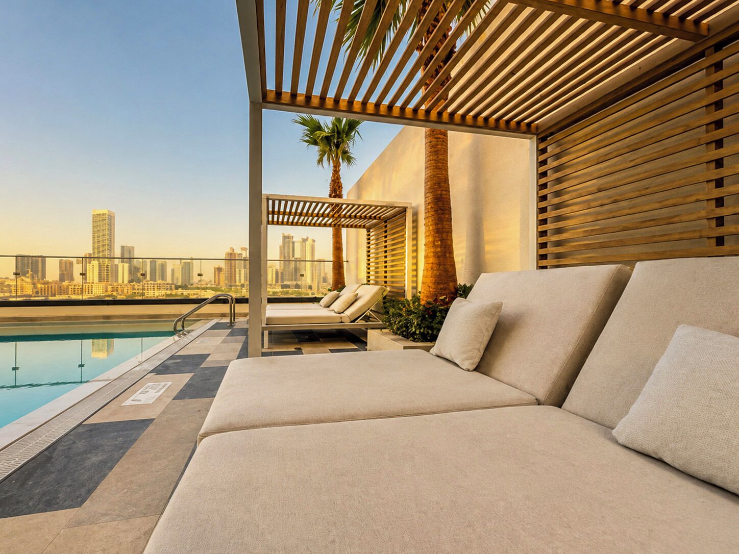 A rooftop pool area with a sofa and lounge chairs, a palm tree, and a city skyline view.