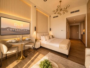 A modern hotel room with a bed, bedside table, and a TV displaying 'DvH COLLECTION'. - Modern Chic Studio Apartment - JVC Dubai (Dubai)