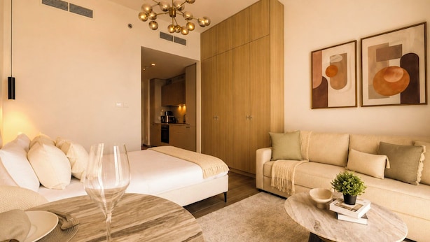 A modern living room with a sofa, a dining table with a glass top, and a kitchen in the background. - Modern Chic Studio Apartment - JVC Dubai (Dubai)