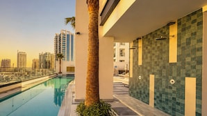 A modern pool area with a clear view of the city skyline, a palm tree, and a tiled wall. - Modern Chic Studio Apartment - JVC Dubai (Dubai)